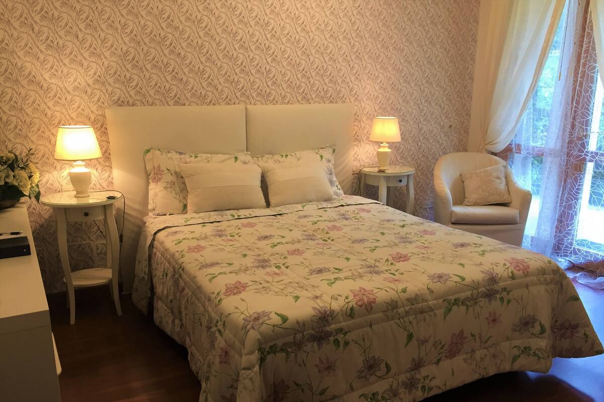 Une chambre à coucher avec une couvre-lit à motifs floraux, deux lampes de chevet, un fauteuil et un papier peint à motifs.