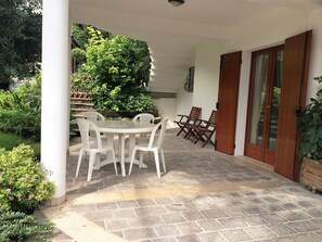 Un patio couvert avec un sol en pierre, des chaises en plastique blanches et une porte en bois.