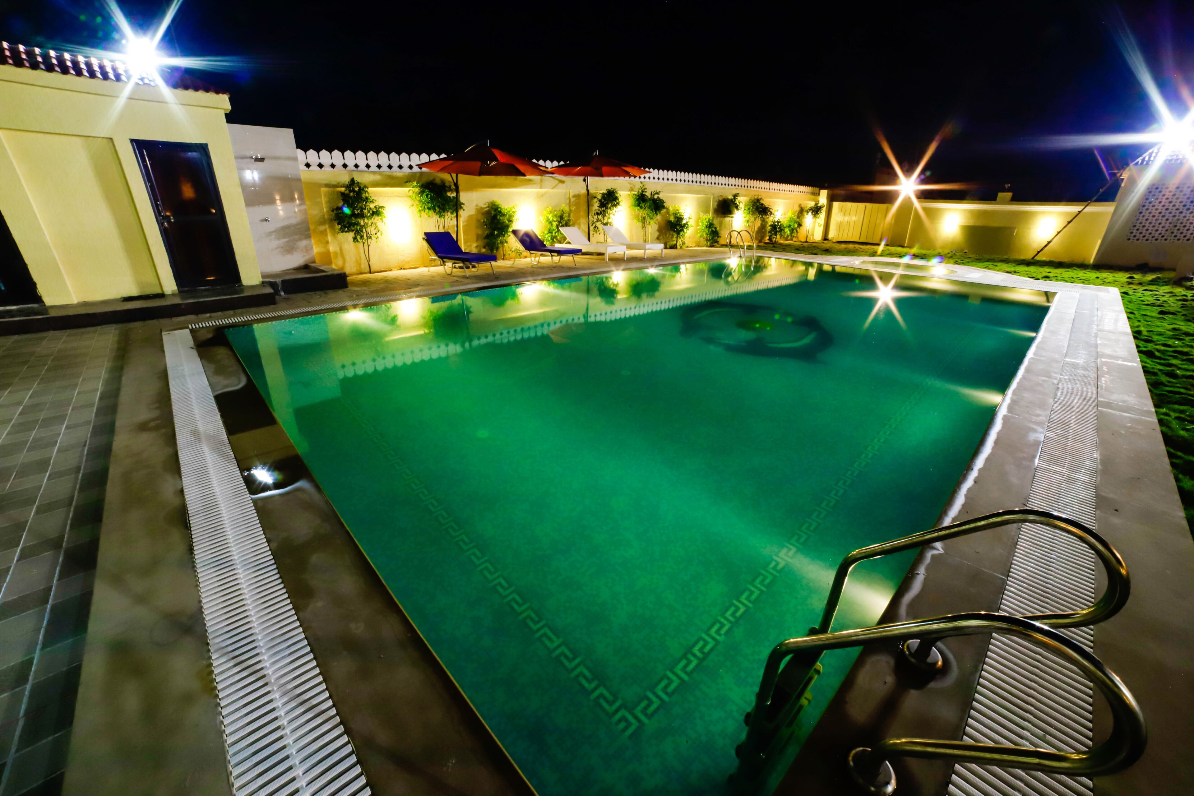 A swimming pool with a diving board, surrounded by lounge chairs and umbrellas, and a white fence in the background.