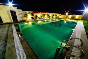 A swimming pool with a diving board, surrounded by lounge chairs and umbrellas, and a white fence in the background.