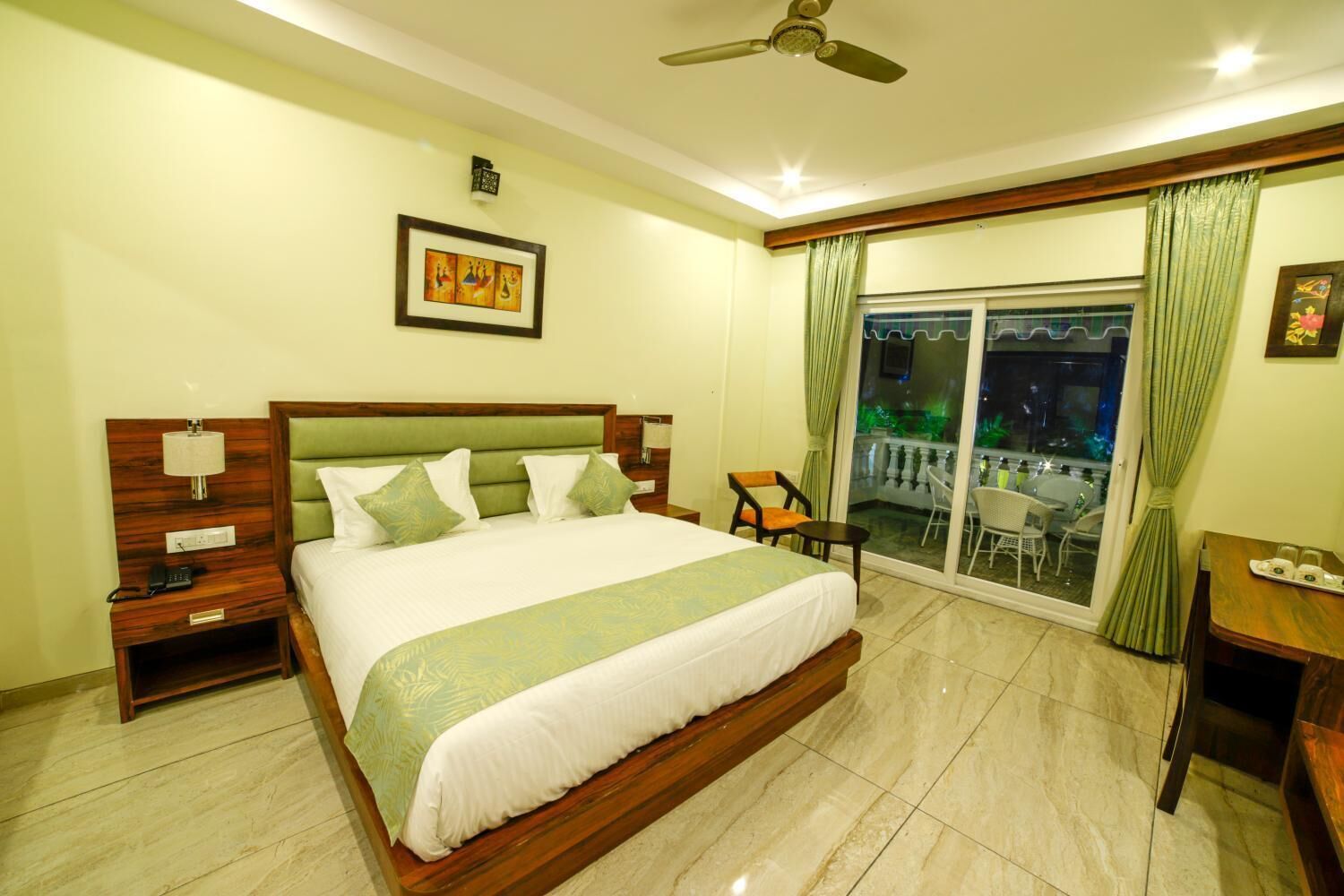 A hotel room with a large bed, a nightstand, a desk, a chair, and a balcony with a view.