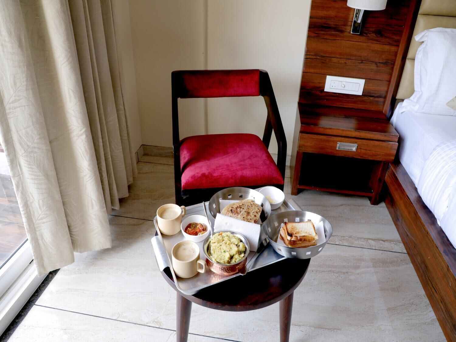 Une chambre d’hôtel avec une table à manger dressée, où l’on voit de la nourriture et des boissons, une chaise rouge, une table de chevet en bois et un lit recouvert de draps blancs.