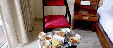 Une chambre d’hôtel avec une table à manger dressée, où l’on voit de la nourriture et des boissons, une chaise rouge, une table de chevet en bois et un lit recouvert de draps blancs.