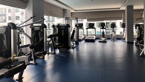 A gym with various exercise machines, large windows, and a blue floor. - Arcadia Family Suite (Pattaya)