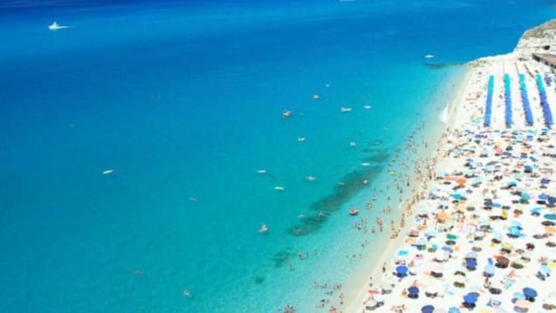 An aerial view of a beach with clear turquoise waters, a sandy shore, and numerous umbrellas.