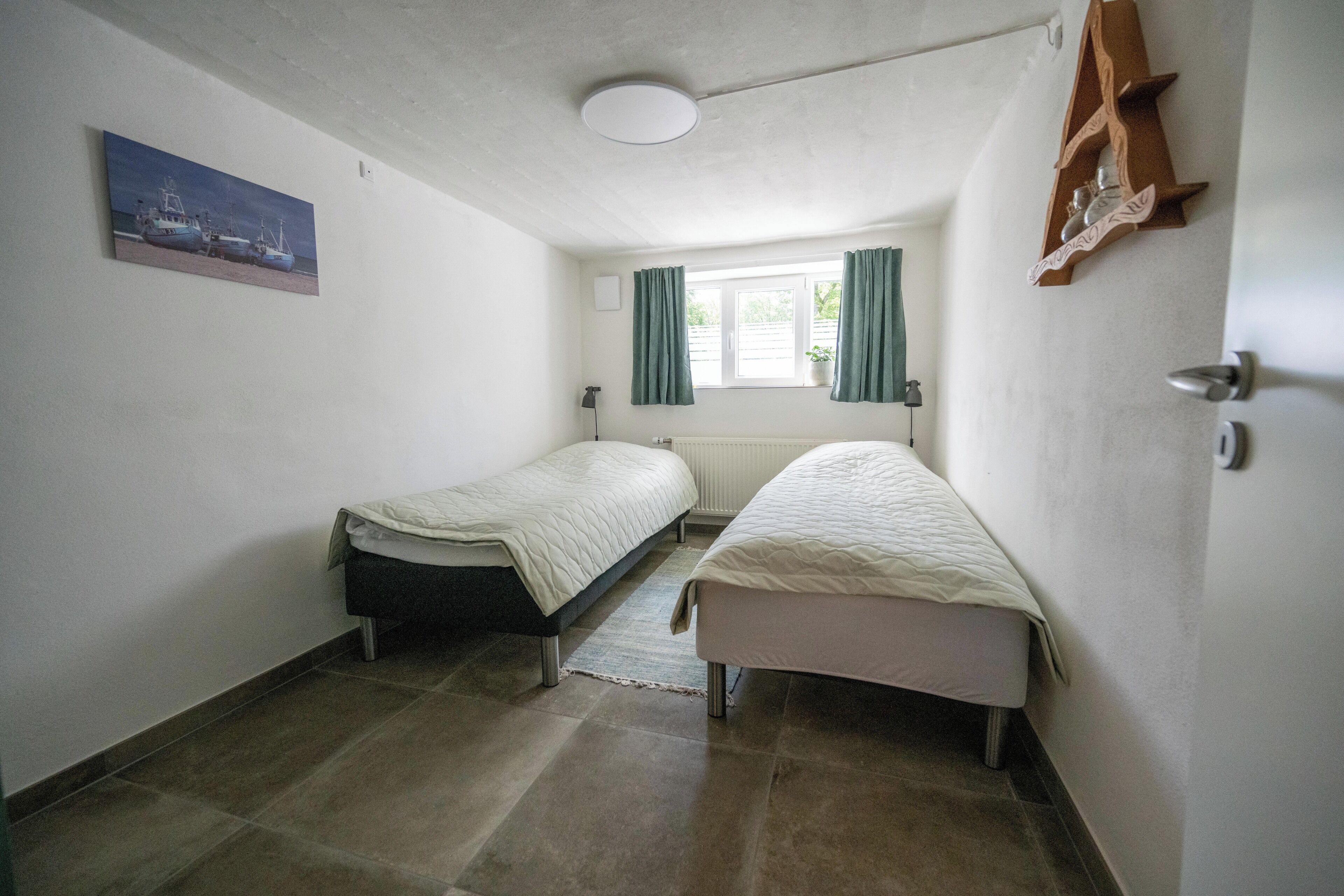 A small, well-lit room with two single beds, a window with curtains, a radiator, and a painting on the wall.