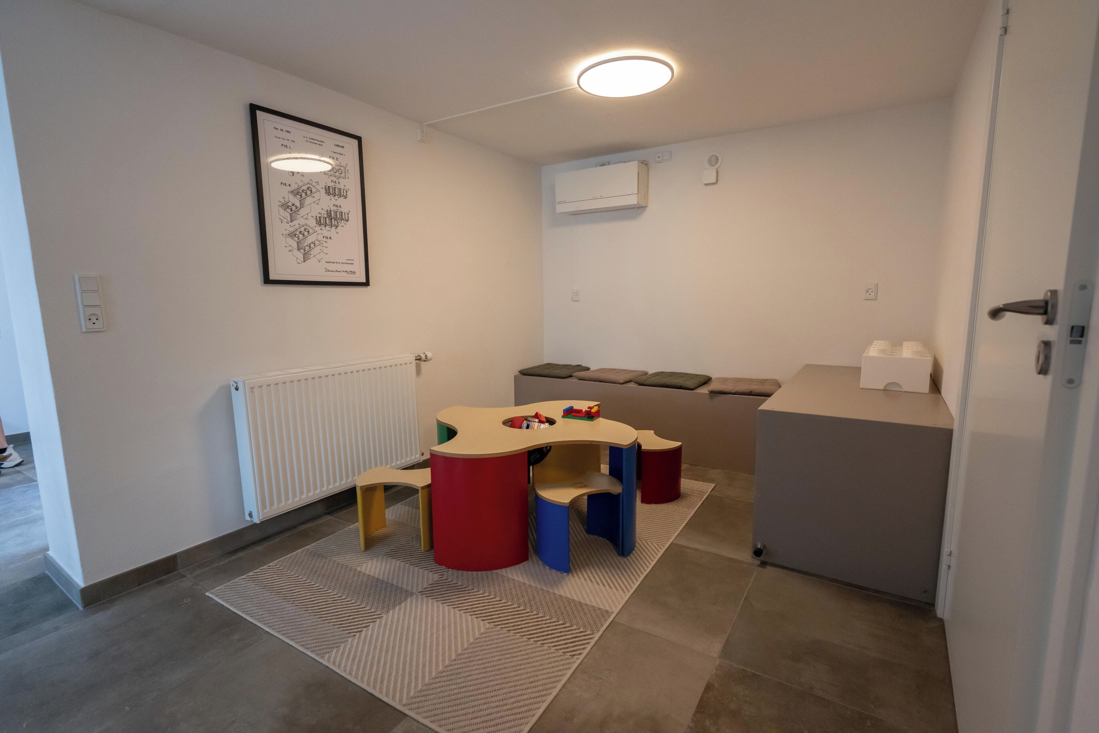 A child's play area with a table and colorful stools, a radiator, a framed picture on the wall, and a small refrigerator.
