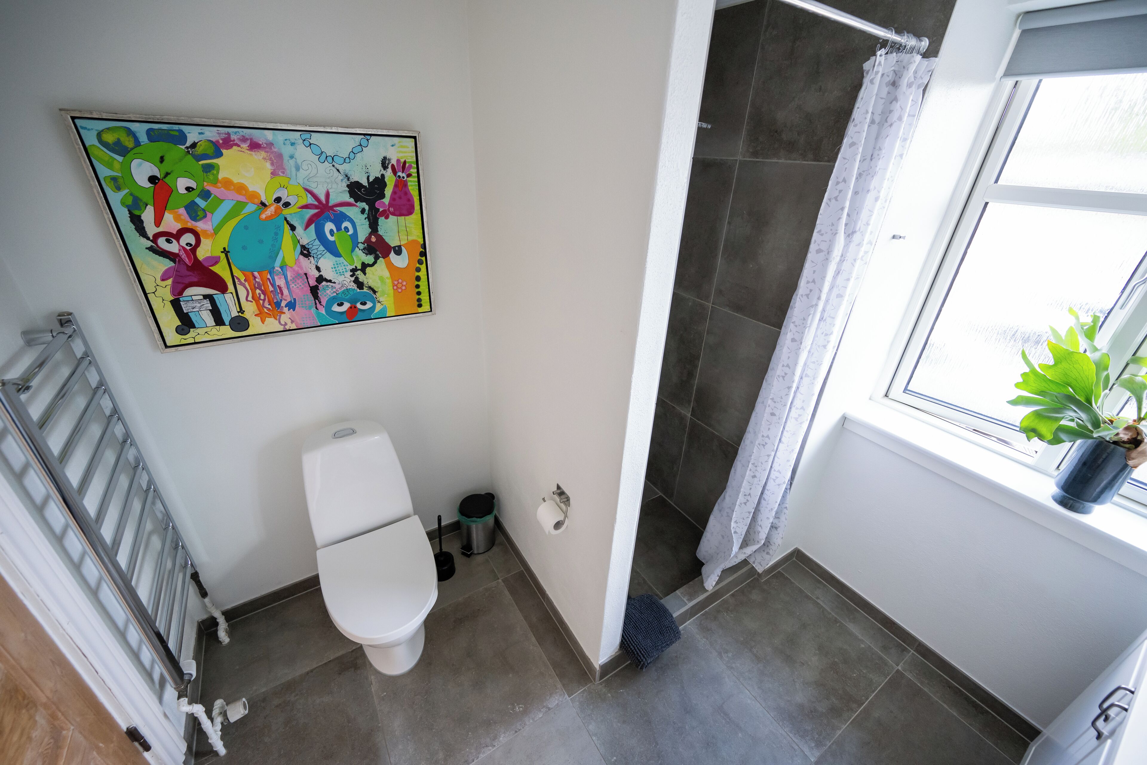 Une salle de bain avec des toilettes, une douche et une fenêtre agrémentée d’une plante.