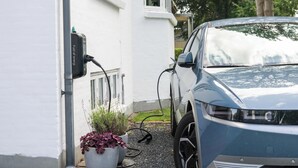 Un automóvil estacionado junto a una estación de carga, un edificio blanco y una planta en maceta.