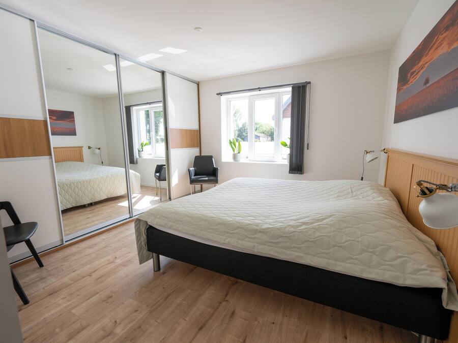 A bedroom with a bed, a chair, a desk, a window with curtains, and a mirror.