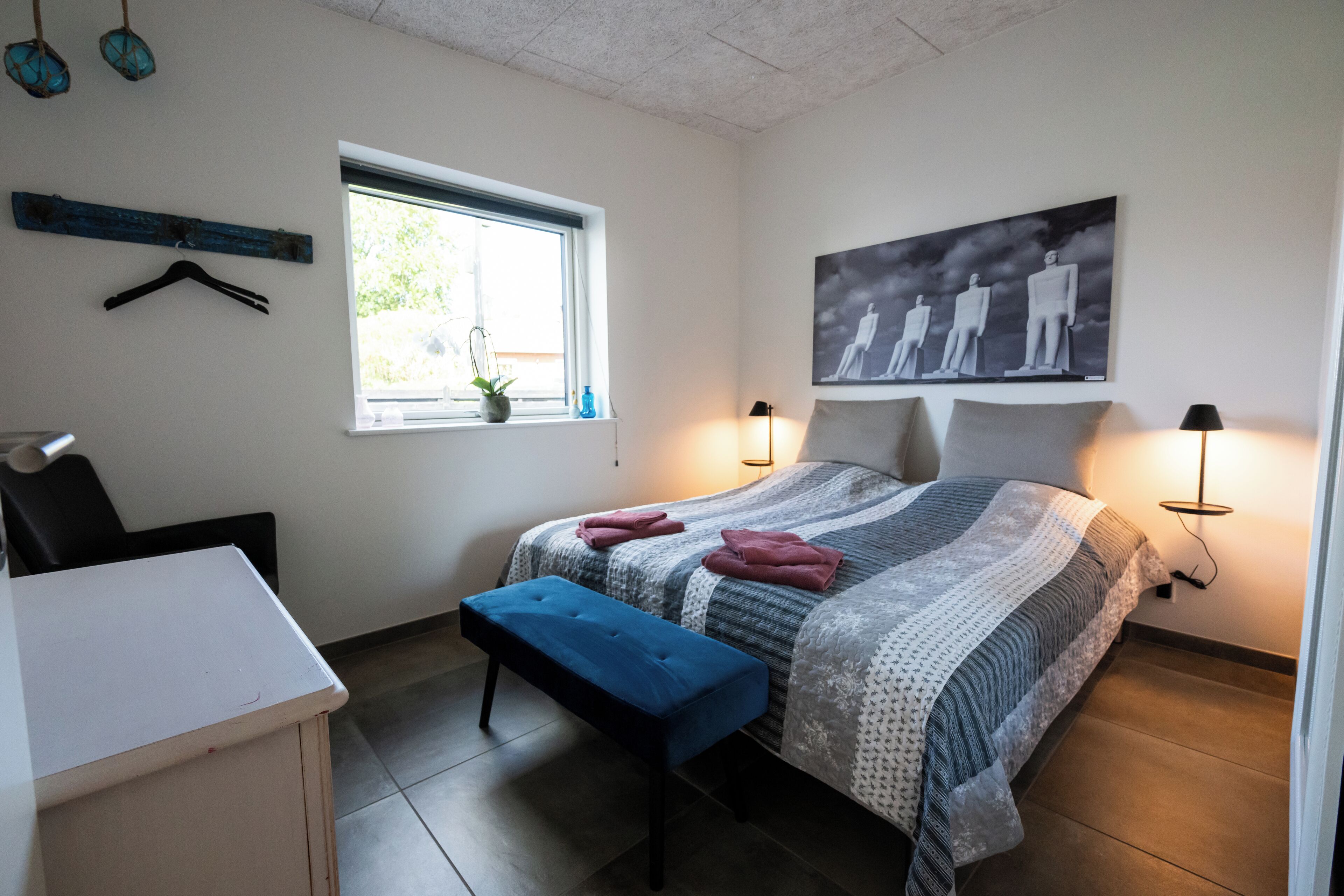 A bedroom with a bed, bench, desk, window, and artwork.