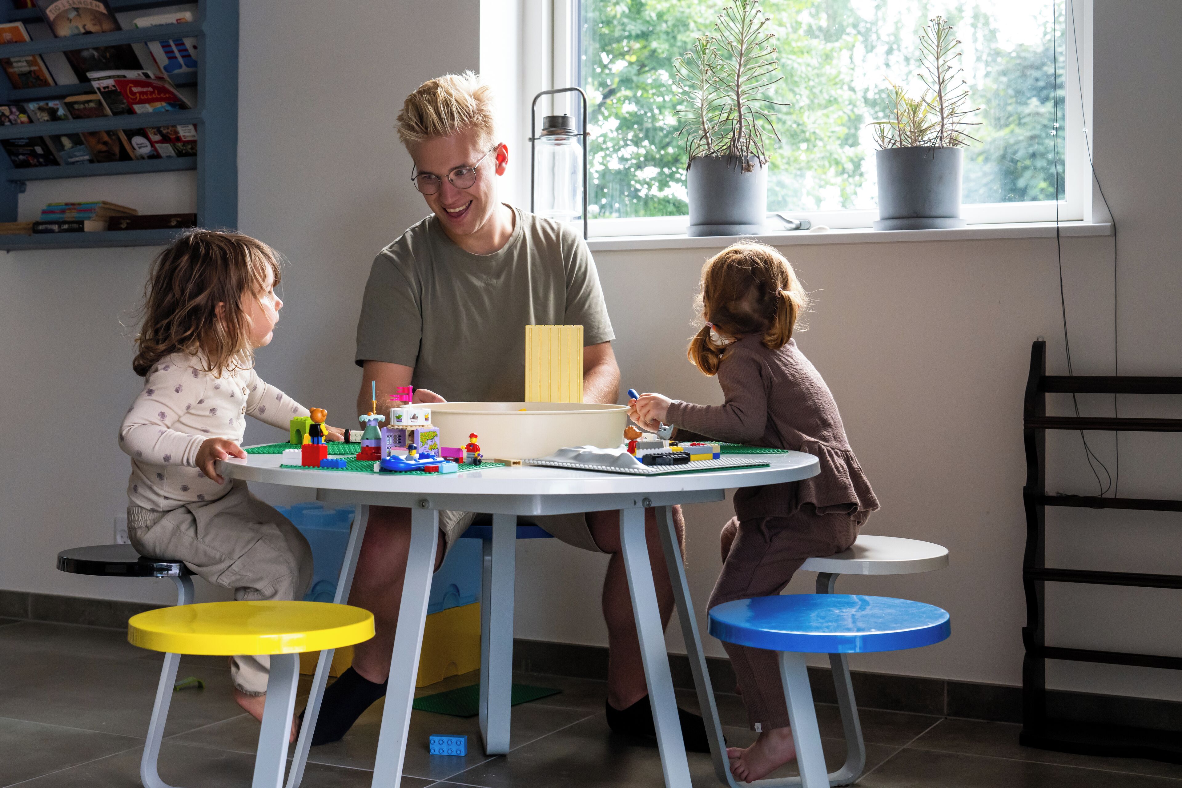 Ein Zimmer mit einem Tisch, an dem zwei Kinder und ein Erwachsener mit Spielzeug spielen. Auf einem Fensterbrett stehen Blumentöpfe und im Hintergrund steht ein Bücherregal.