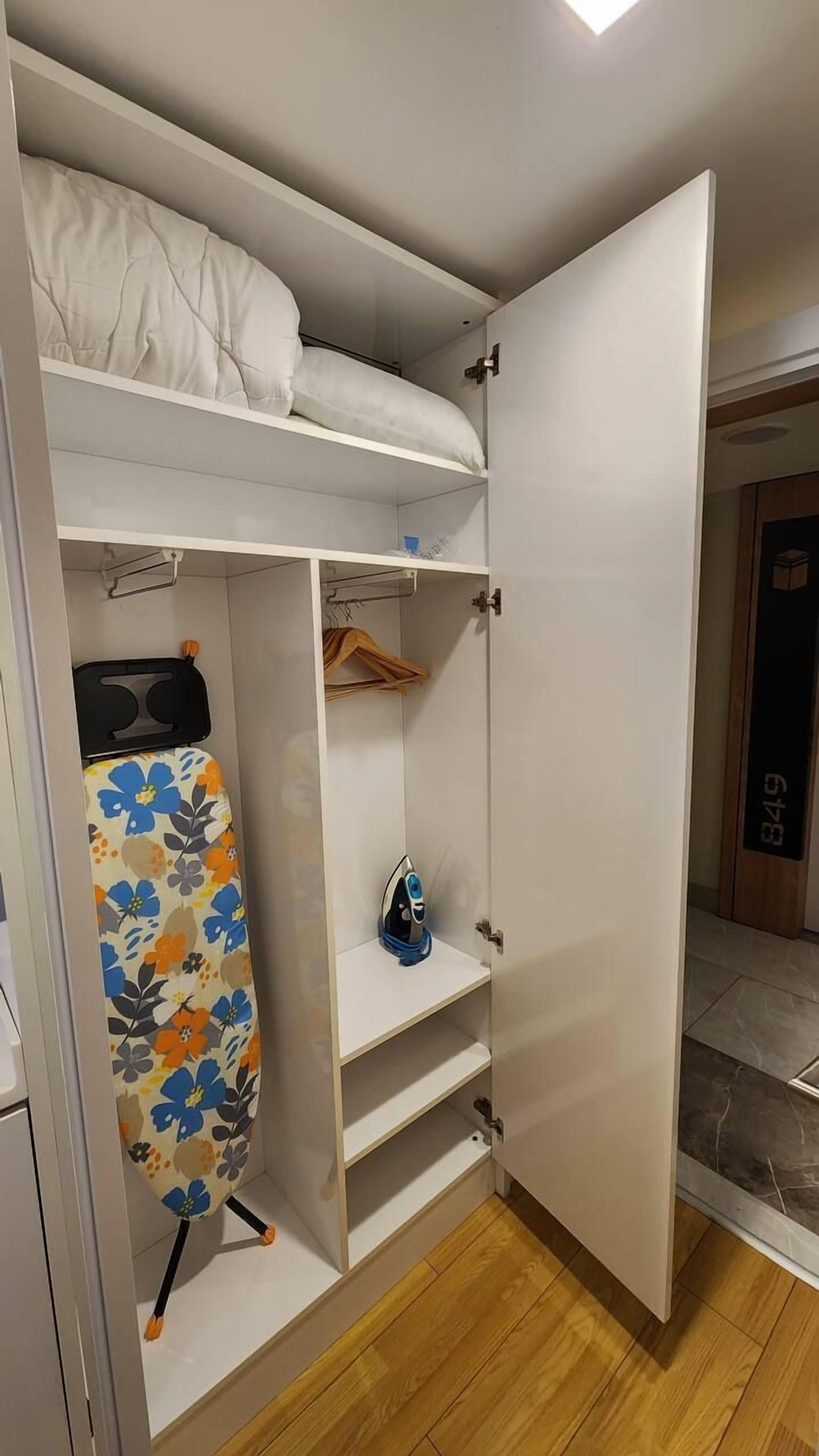 A walk-in closet with a bed above, featuring a hanging rack, shelves, and a colorful ironing board.