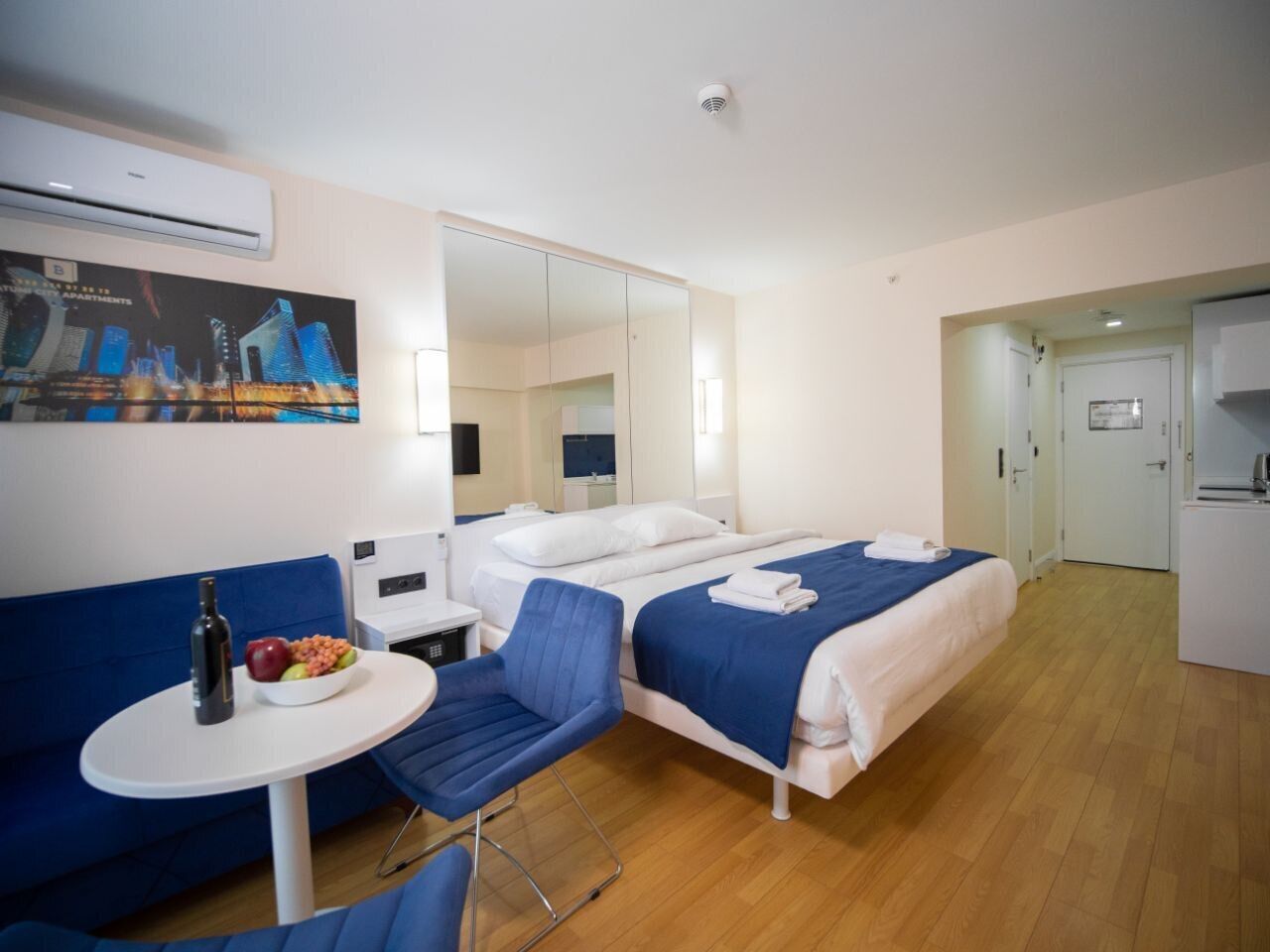 Une chambre d’hôtel avec un lit, un bureau, une chaise, une petite table avec une bouteille et des fruits, et un miroir.