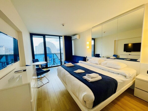 A hotel room with a large bed, a desk with a chair, a TV, a balcony with a view of the city, and a large mirror.