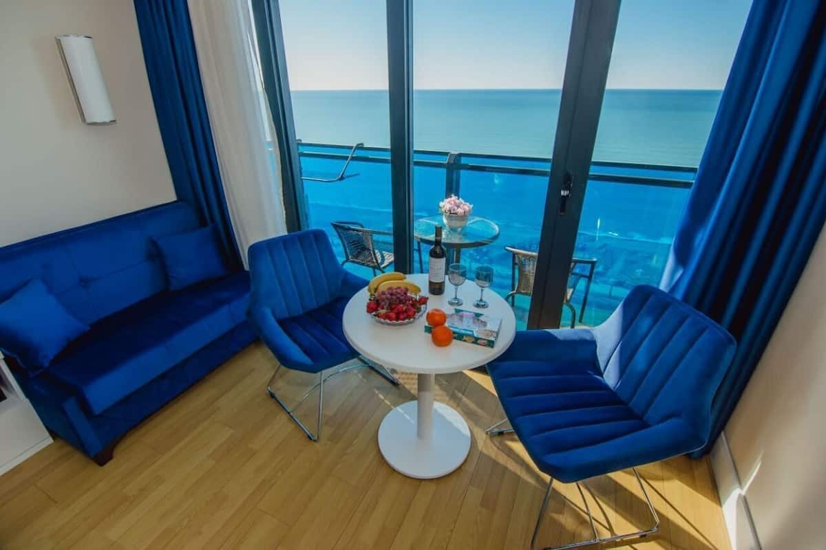 Een balkon met een blauwe bank, twee blauwe stoelen, een kleine ronde tafel met een fles, glazen en fruit.