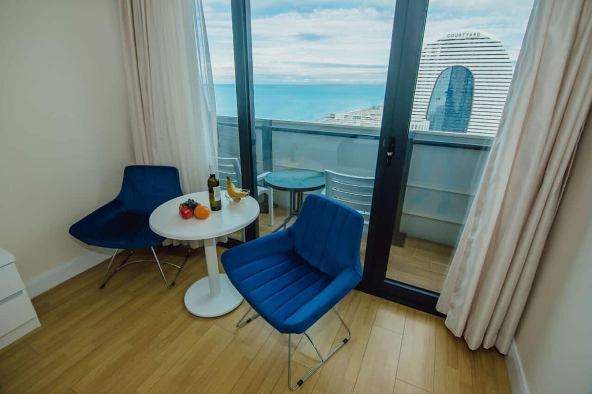 Een balkon met uitzicht op zee en een gebouw met de tekst 'COURTYARD'. Er staan twee blauwe stoelen, een klein rond tafeltje met fruit en flessen, en een witte stoel.