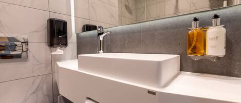 A modern bathroom with a white sink, a handheld soap dispenser, and two bottles of LAPE body wash on a gray-tiled wall.