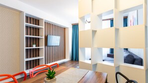 Un salón moderno con suelo de madera, una mesa de comedor con sillas, un televisor montado en una pared de paneles de madera y una estantería con una planta.