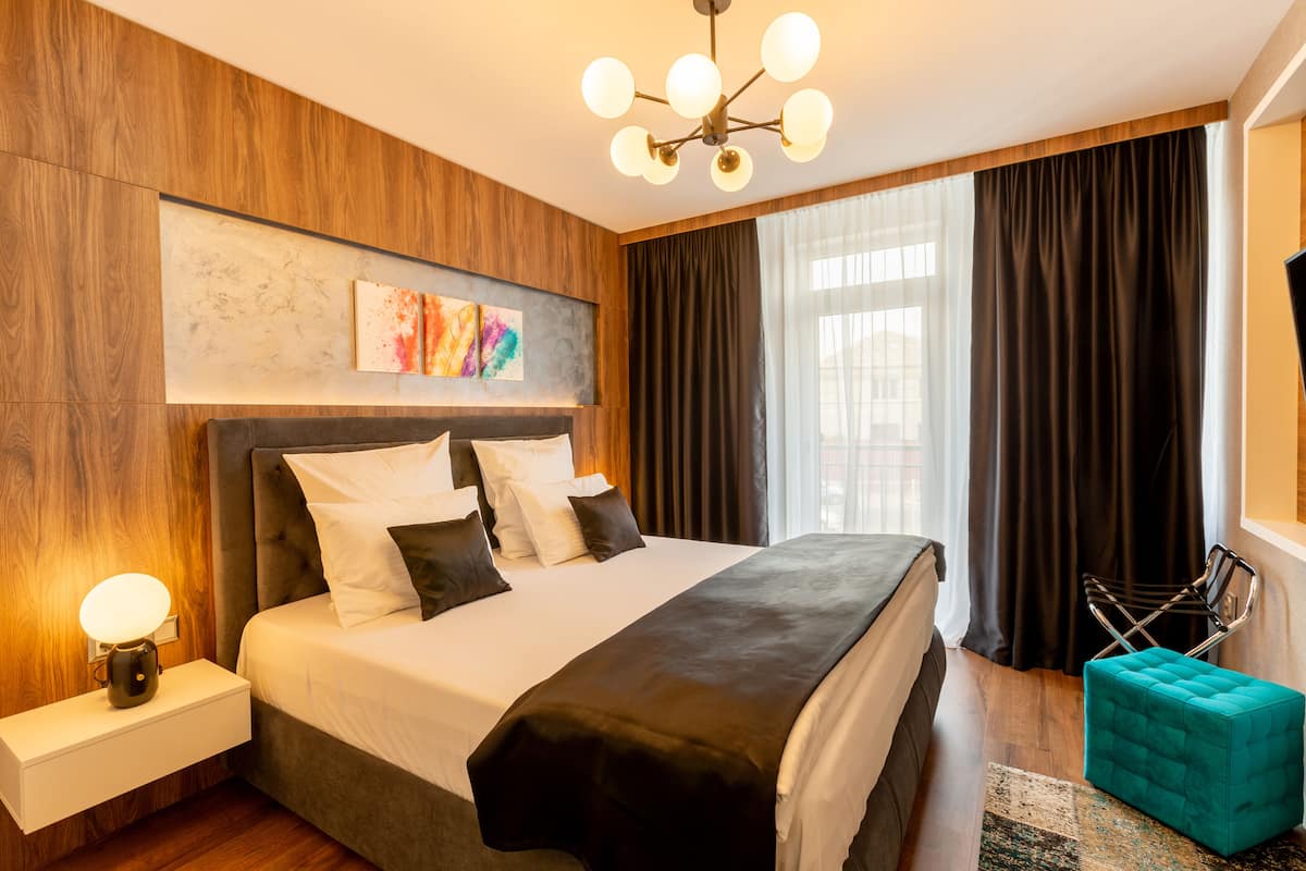 Una habitación de hotel moderna con una cama grande, una mesita de noche con lámpara, una obra de arte en la pared y vistas a la ciudad a través de amplios ventanales.