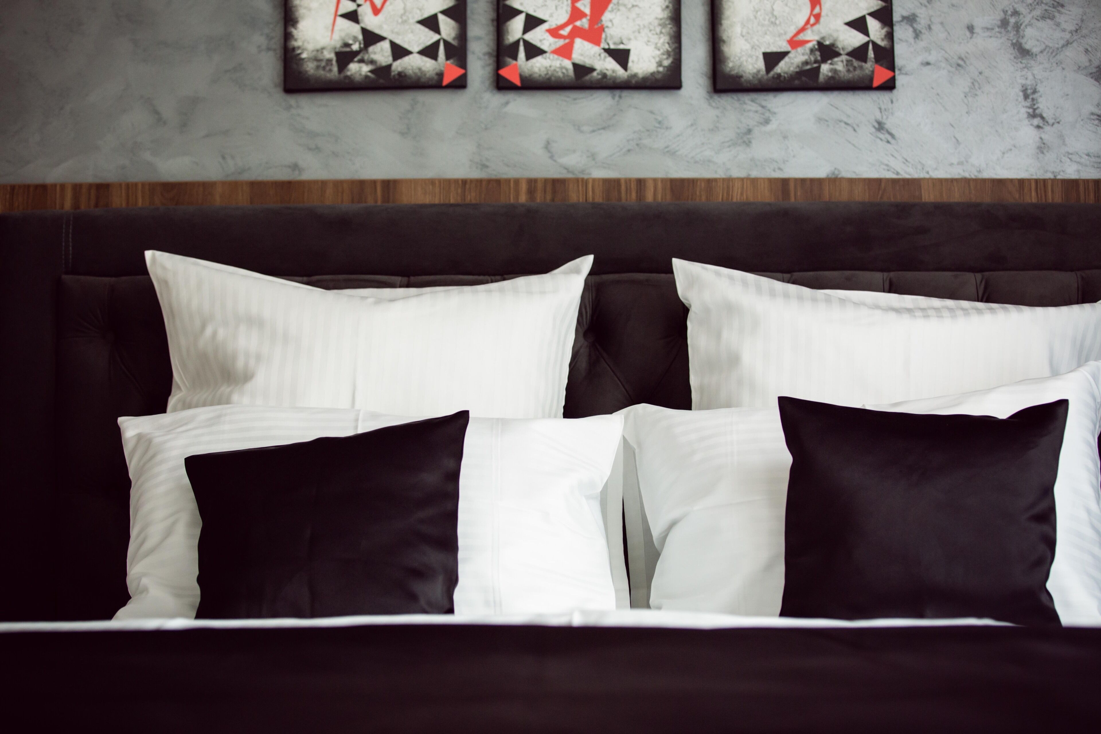 Un lit bien fait, avec des oreillers blancs et noirs, une tête de lit sombre et trois tableaux abstraits accrochés au mur au-dessus.