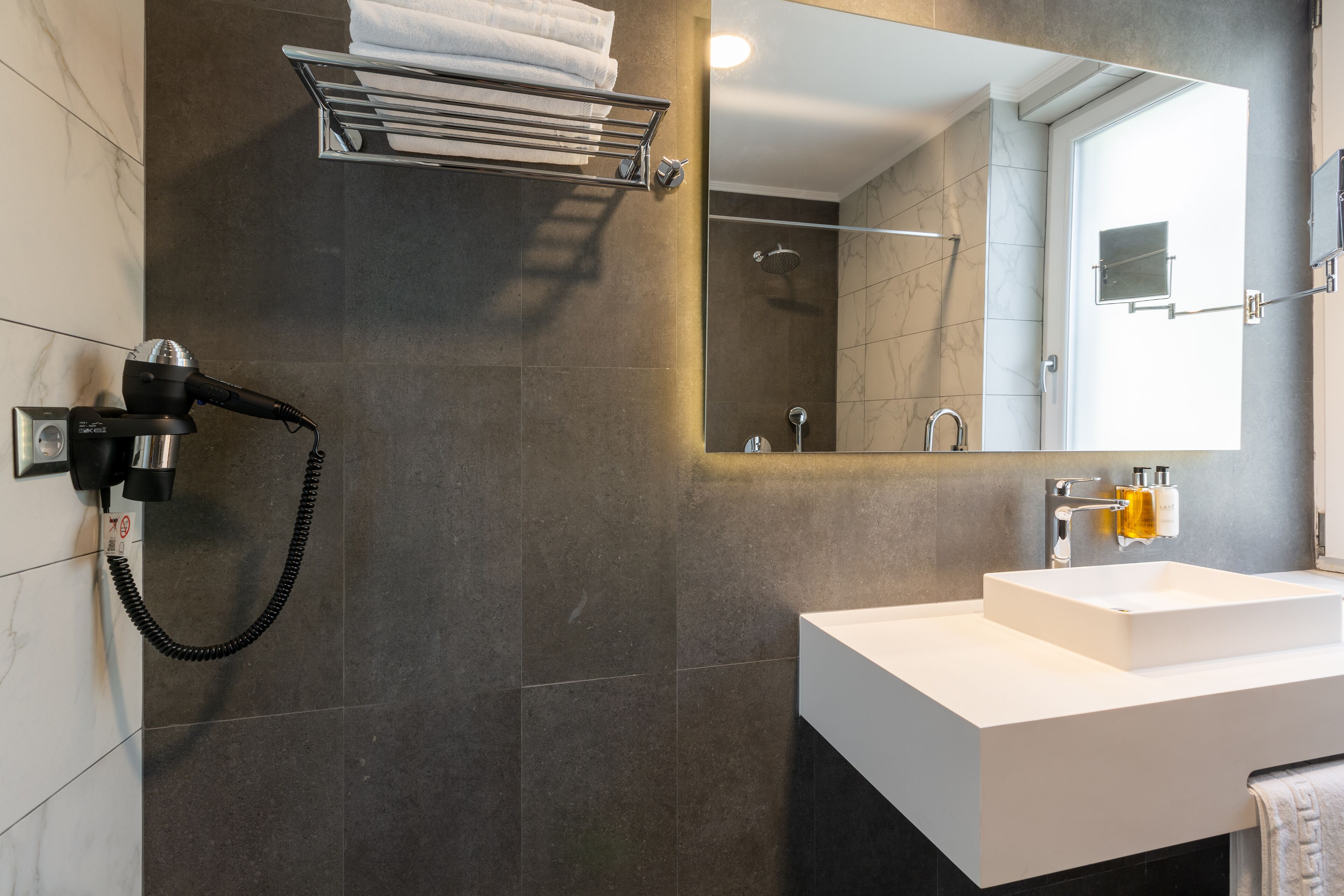 A modern bathroom with a white sink, a towel rack, a hairdryer, and a shower area.