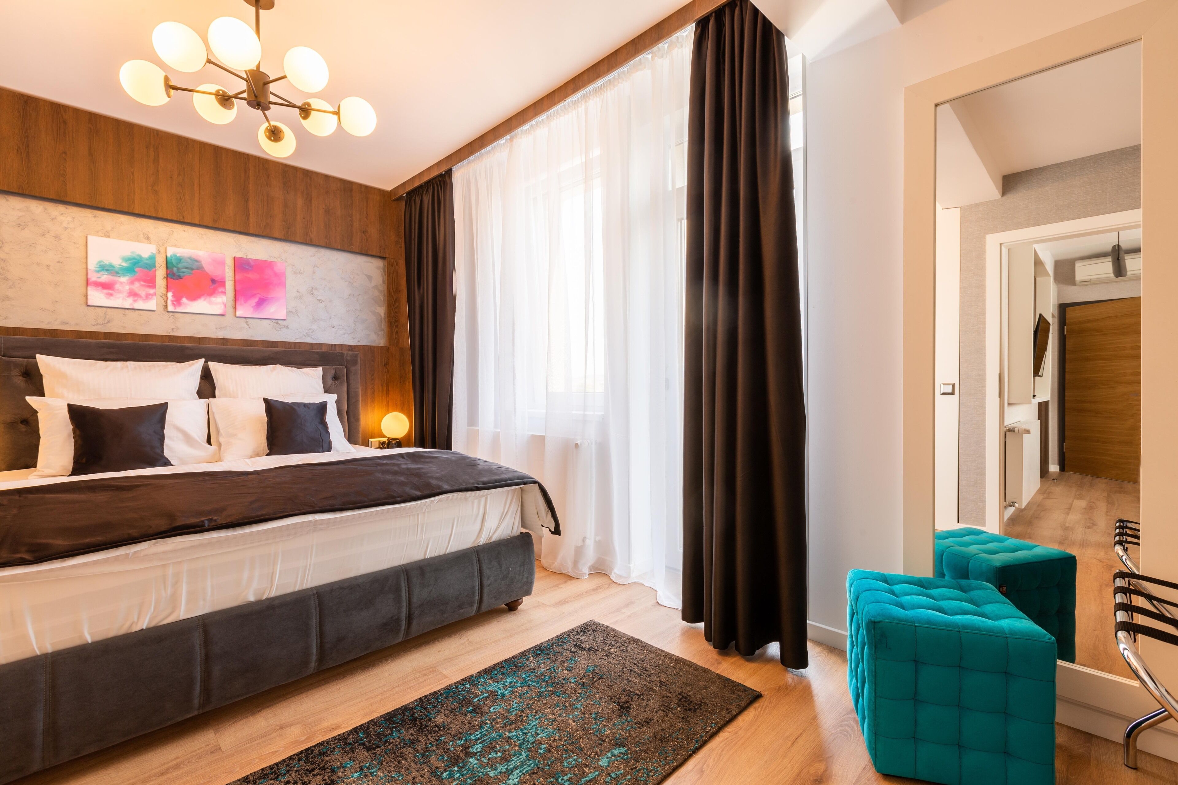 Una camera da letto moderna con un letto grande, una testiera decorata con opere d'arte, una lampada da comodino, una finestra con tende leggere, un tappeto scuro e un pouf color acquamarina.