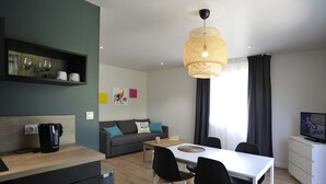 Una sala de estar moderna con sofá, mesa de centro, mesa de comedor y sillas. Hay un televisor sobre un mueble bajo, una cafetera y una estantería con objetos decorativos.