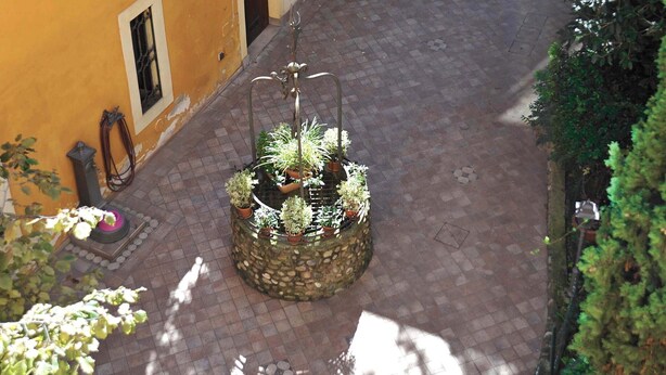 Ein Innenhof mit gefliestem Boden, ein kleiner Garten mit Blumentöpfen und ein gelbes Gebäude mit grünen Fensterläden.
