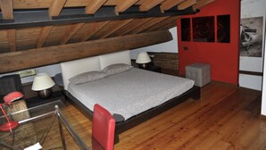 Ein Schlafzimmer mit Holzboden, einem Bett mit grauem Topper, zwei Nachttischlampen und einer roten Wand mit einem Bild.