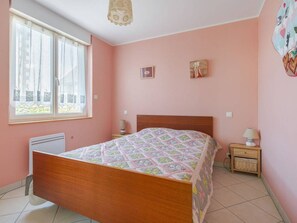 A bedroom with a wooden bed, a window with curtains, a bedside table with a lamp, and a wall with two framed pictures.