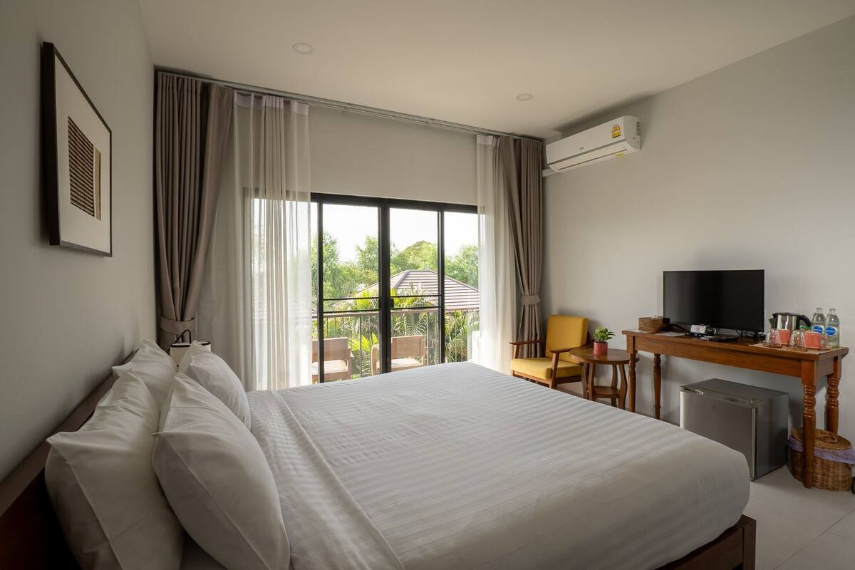 A hotel room with a large bed, a desk with a TV, a small fridge, and a view of greenery outside.