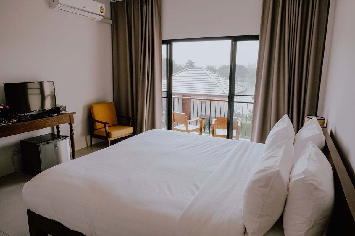 A hotel room with a large bed, a desk with a television, and a balcony with chairs.