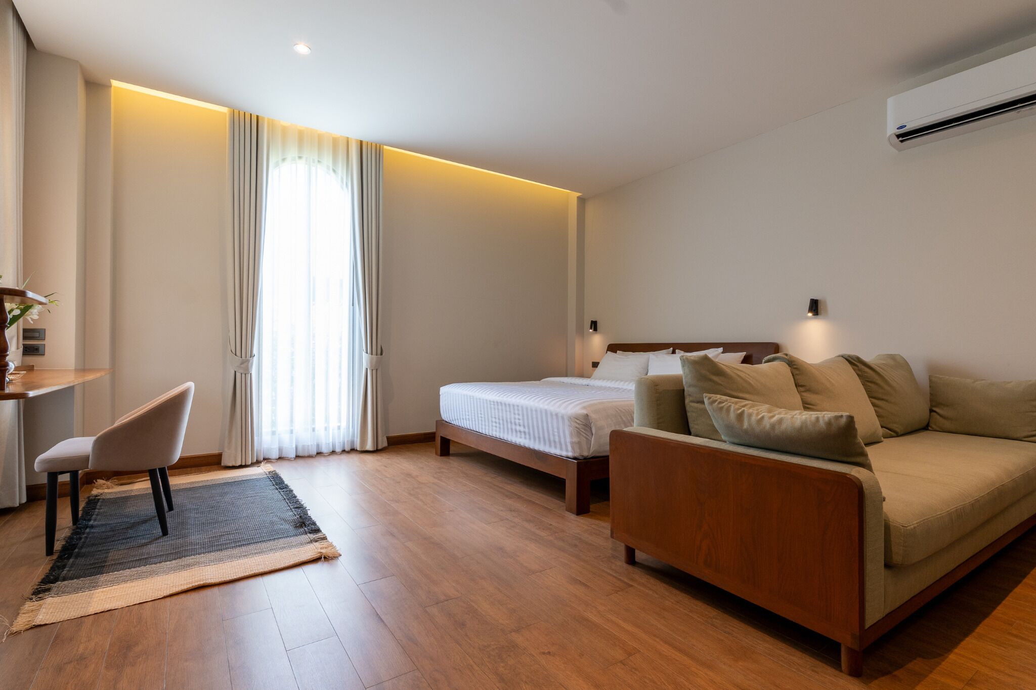 A modern hotel room with a wooden floor, a bed, a sofa, a desk, and a chair.