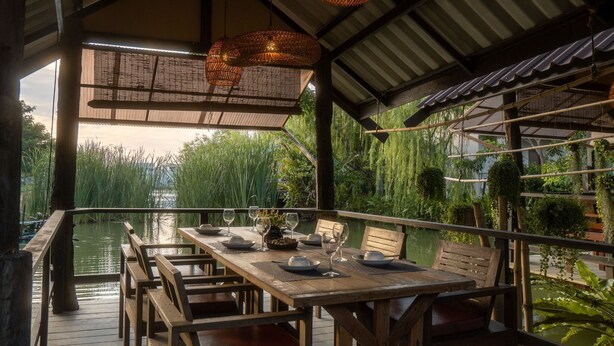 An outdoor dining area with a wooden table and chairs, surrounded by lush greenery and a view of a water body. - Ploen Pirom (Bangkok)