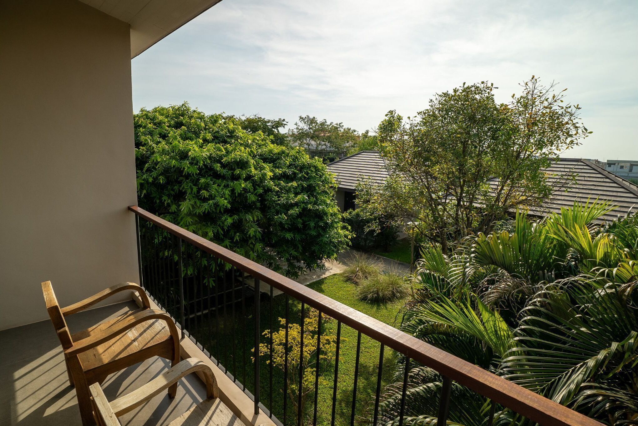 A balcony with a wooden chair, a metal railing, and a view of a garden with various plants and a building in the background.
