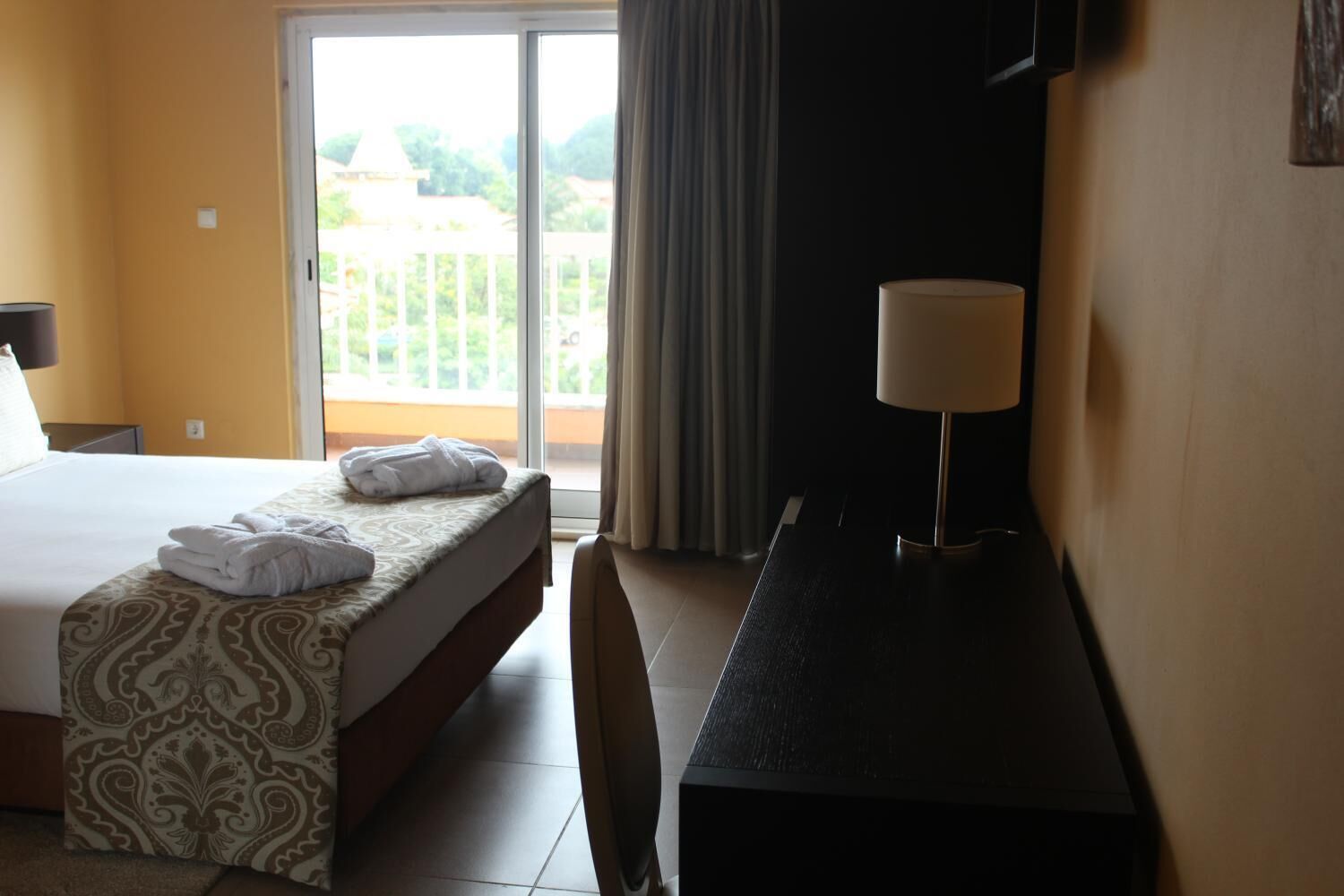 A hotel room with a bed, a desk, a lamp, and a view of the outdoors through a sliding door.