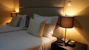 Habitación de hotel con dos camas, cada una con sábanas blancas y numerosas almohadas. Hay lámparas de noche y un teléfono en una mesita.