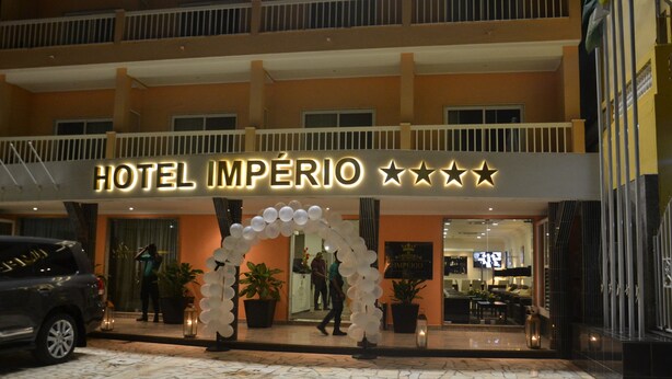 Entrada de hotel con un cartel que dice "HOTEL IMPÉRIO" y cuatro estrellas. Hay plantas en macetas y un arco decorativo blanco en el exterior.