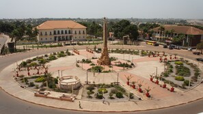 Una plaza pública con un monumento central, zonas verdes circundantes y un edificio con tejado de tejas al fondo.