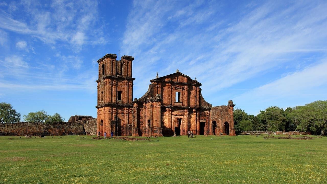 An ancient, red-brick church with a bell tower and a large arched doorway, surrounded by a grassy field and trees.