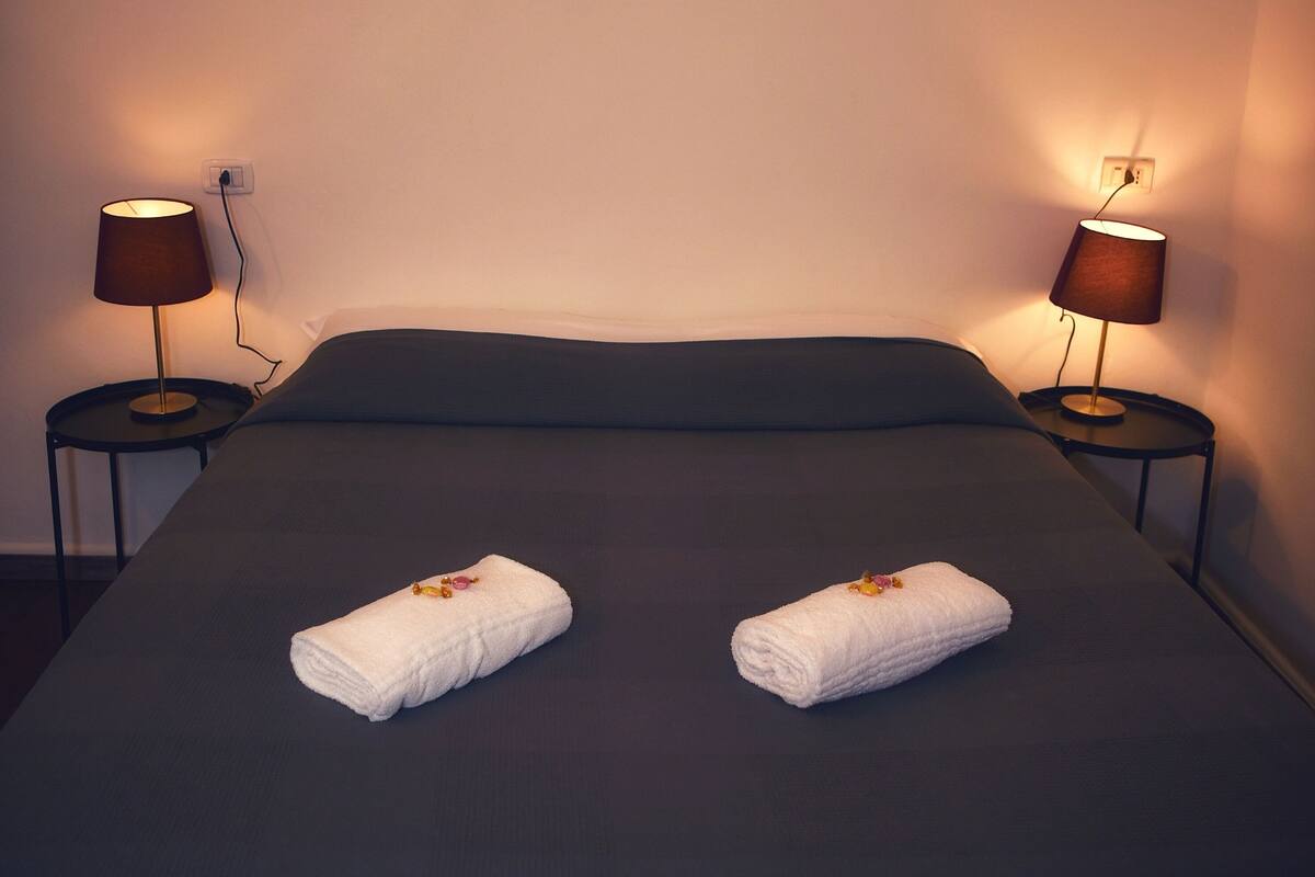 A hotel room with a bed, two bedside lamps, and a cityscape painting on the wall.