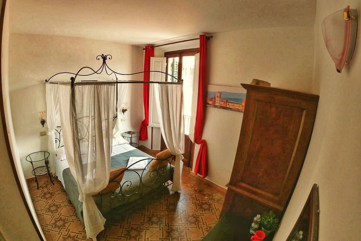 A bedroom with a bed, a small table, a chair, a cabinet, and a view of a building through a window.
