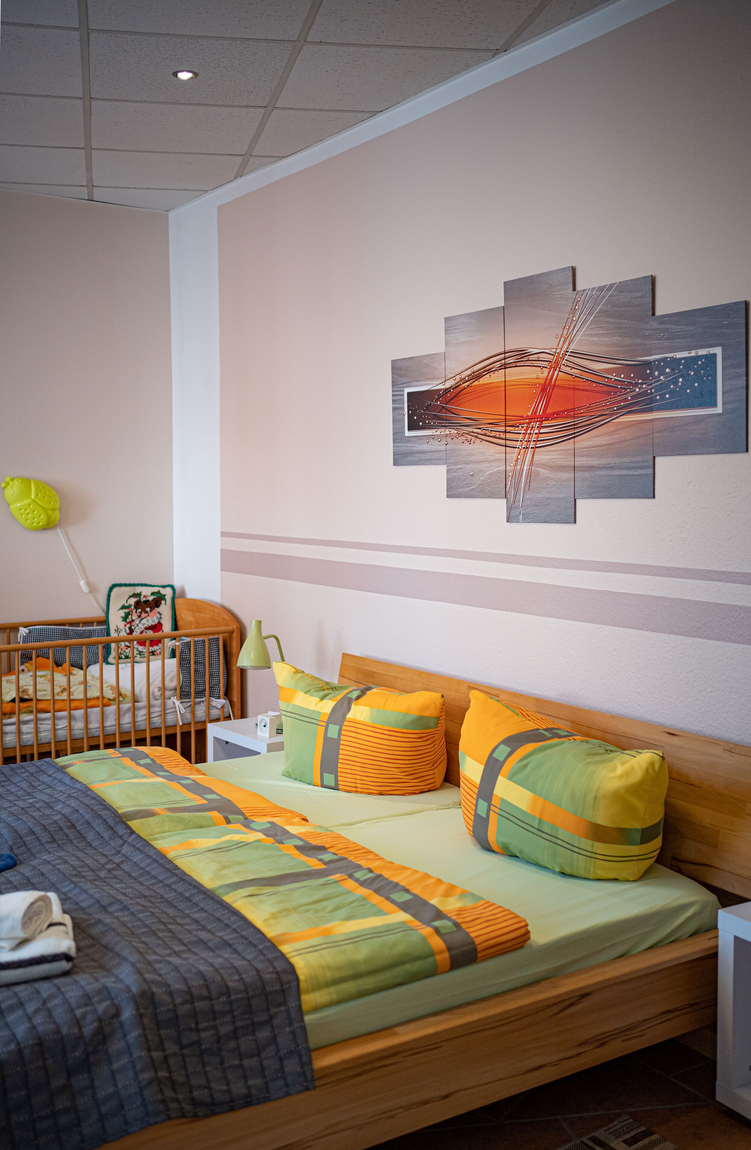 A bedroom with a wooden bed, a crib, a wall-mounted artwork, and a bedside table.