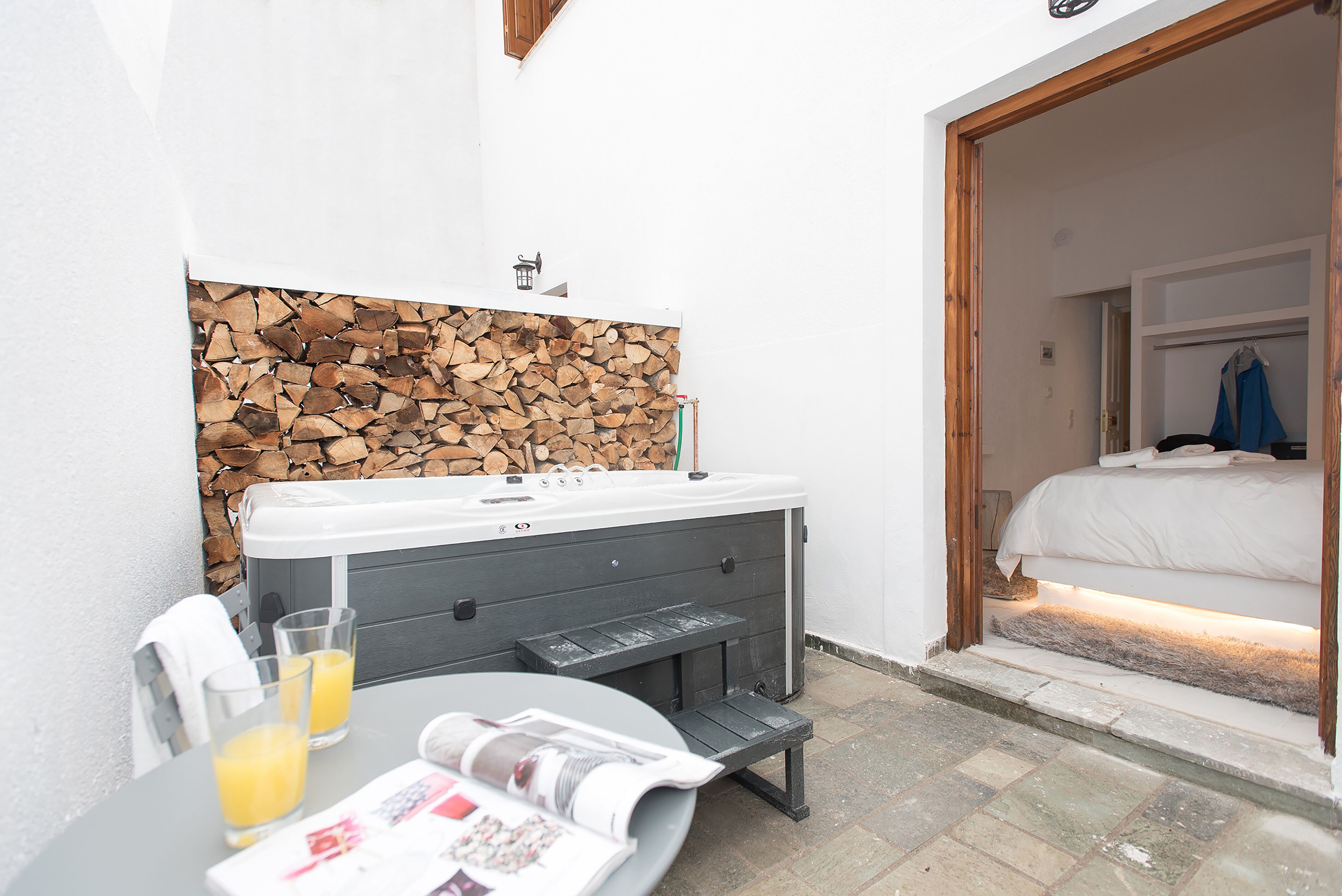 Ein kleiner Sitzbereich im Freien mit einem Tisch, zwei Gläser Orangensaft und einer Zeitung. Im Hintergrund ist eine Sitzecke erkennbar, und durch eine offene Tür ist ein Schlafzimmer mit einem Bett und einem an der Wand hängenden Kittel zu sehen.