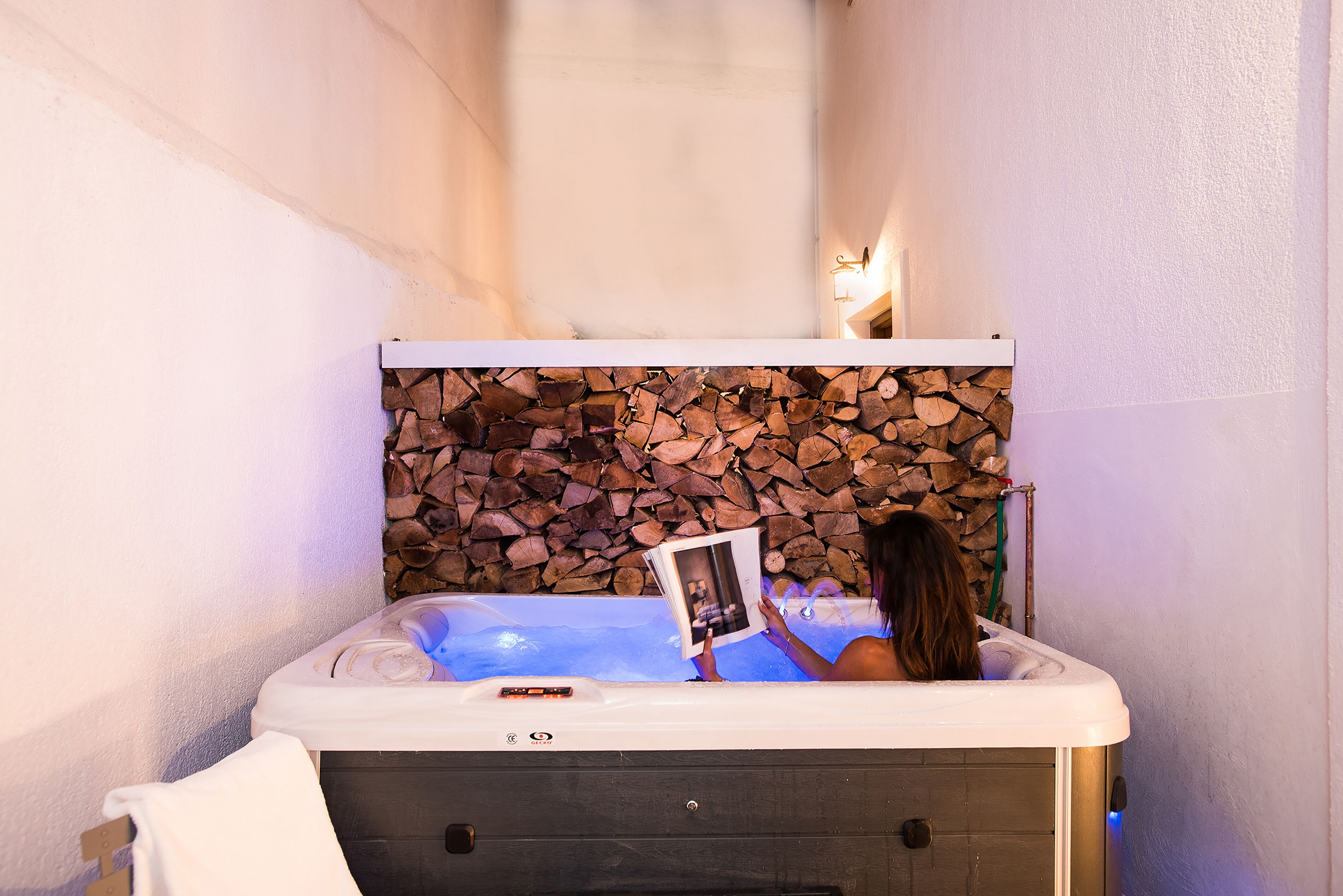 A hot tub with a stone wall backdrop, a towel on a rack, and a person reading a book.