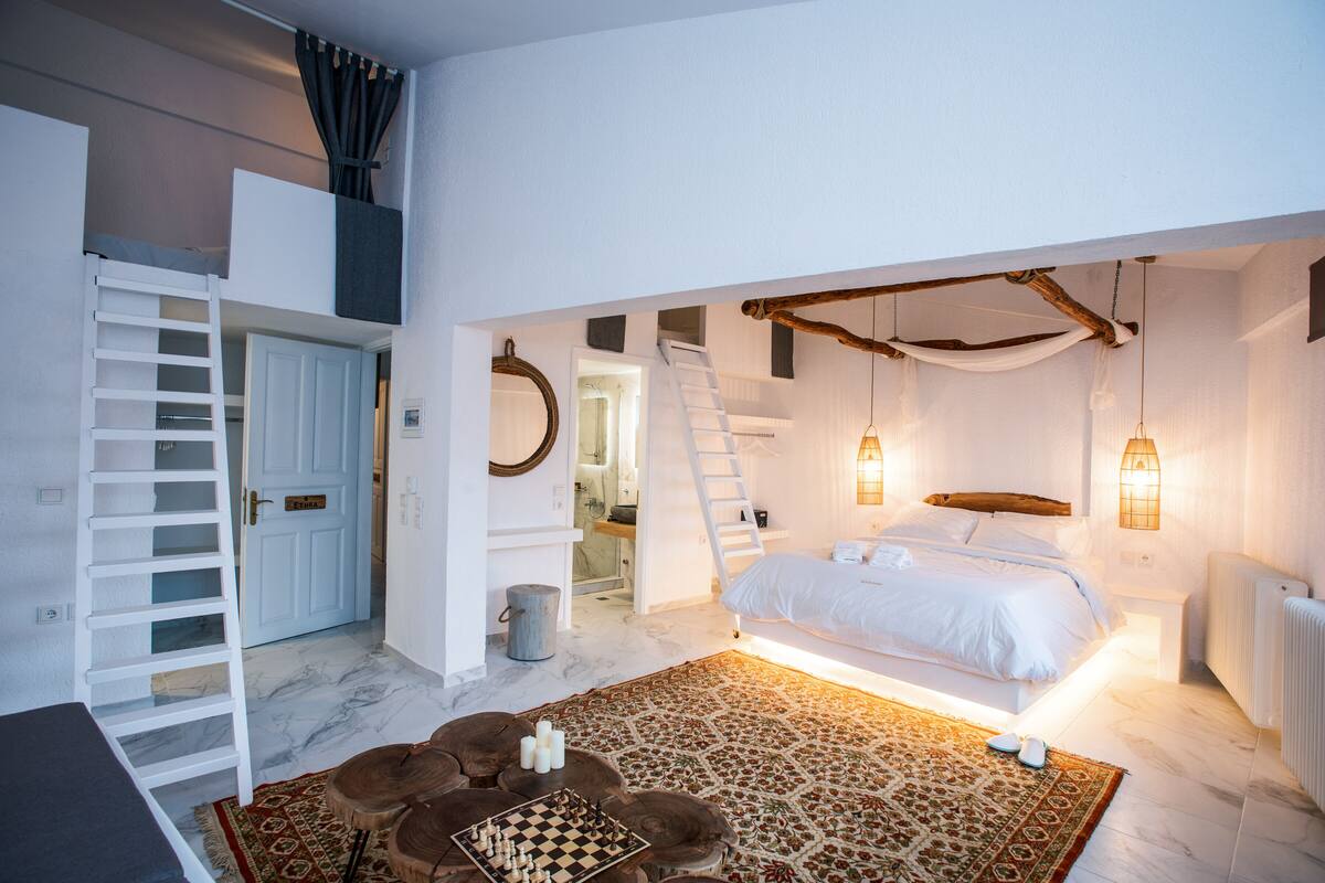 A bedroom with a loft bed, a wooden ladder, a white door, a small table with chess pieces, and a patterned rug.