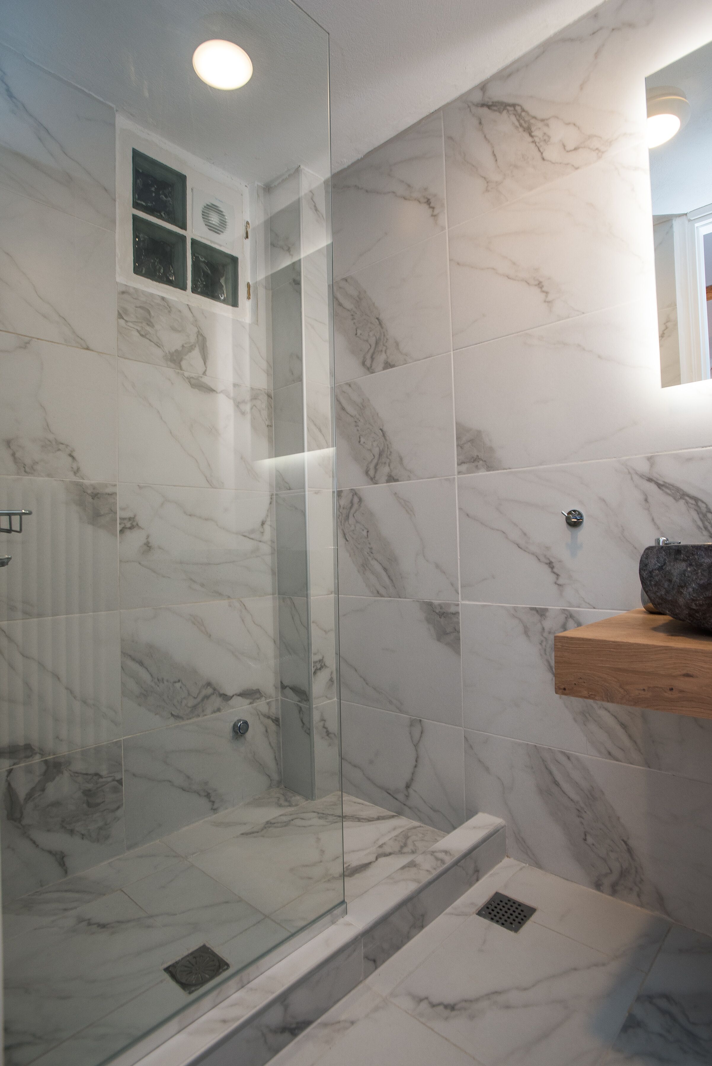 A modern bathroom with marble walls, a glass shower enclosure, and a wooden shelf with a vessel sink.