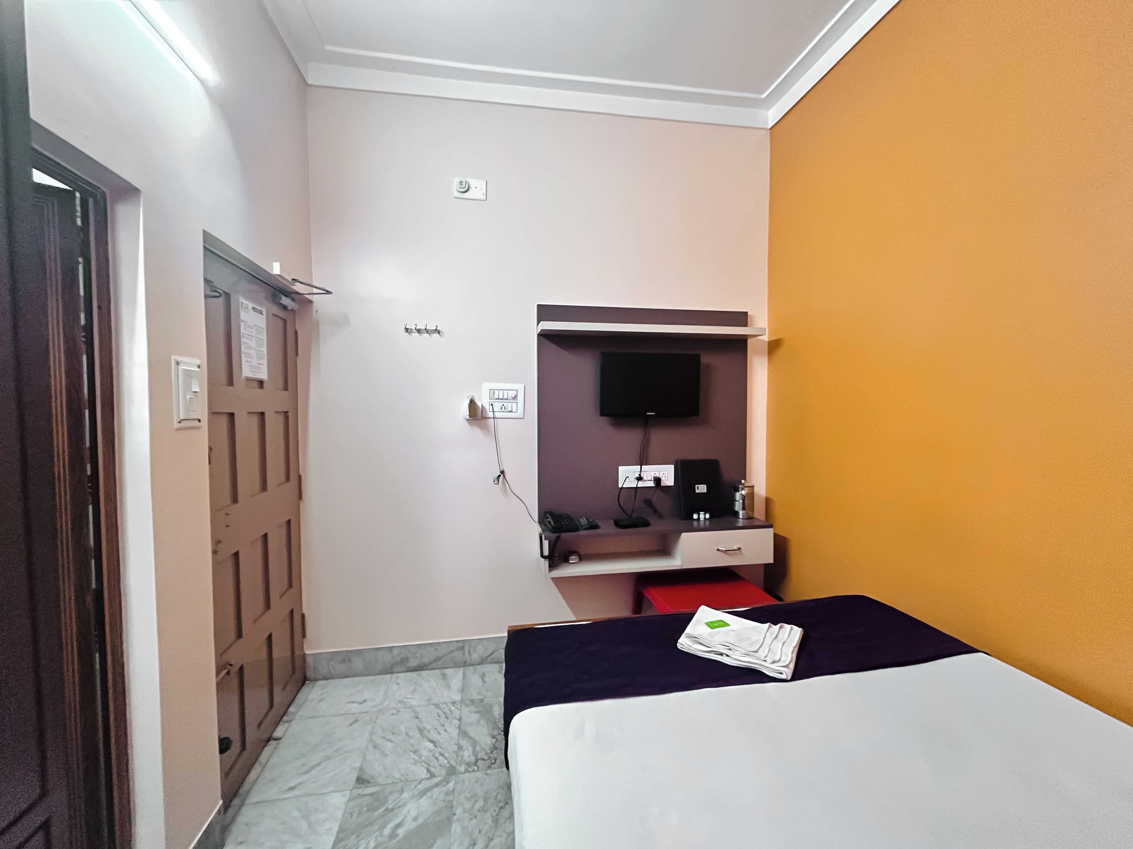 A hotel room with a bed, a desk with a computer, a TV mounted on the wall, and a door.