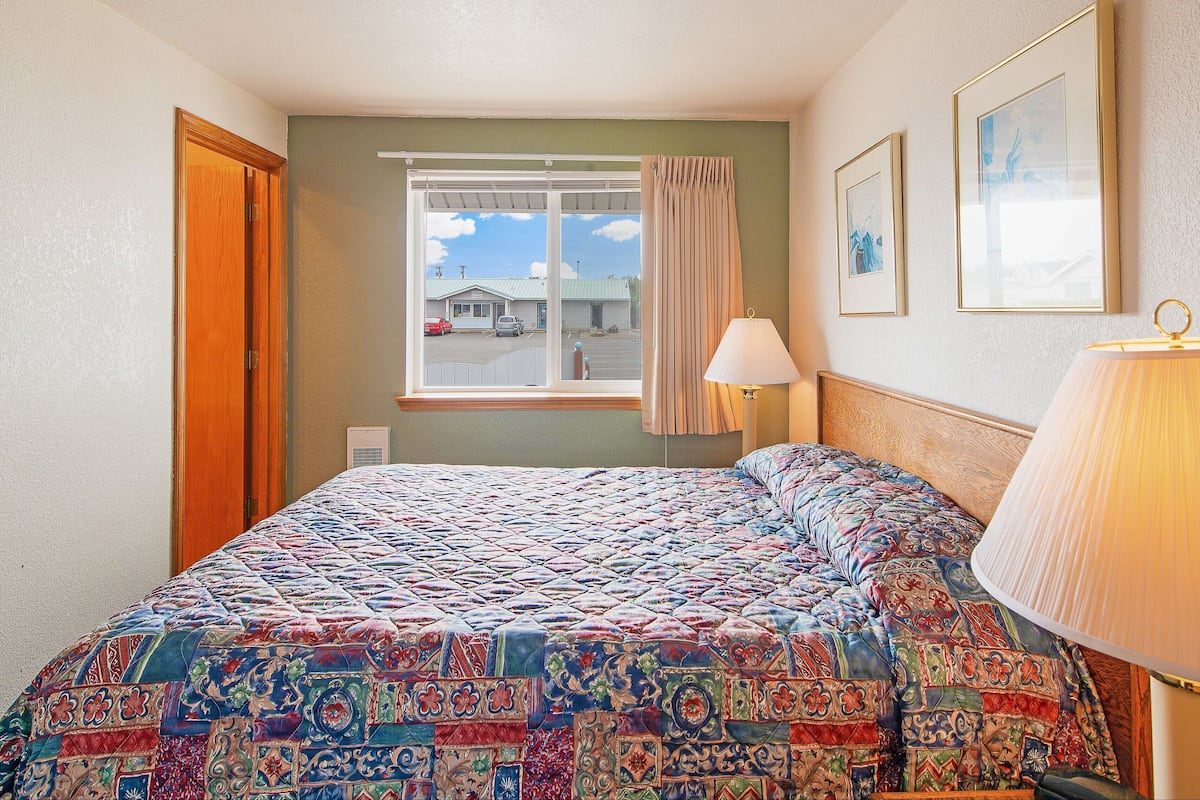 A bedroom with a large bed, a window with curtains, a wall-mounted lamp, and framed pictures.